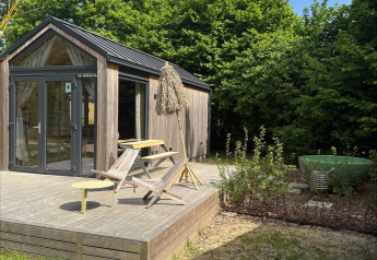 Pequeña casa de madera con terraza, mobiliario exterior y dutchtub, rodeada de árboles en Urban-Gardens, Bélgica.