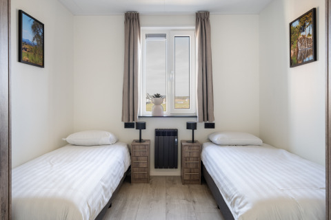 Dos camas individuales en una habitación luminosa y sencilla con ventana, cuadros y mesitas en Zandduin Deluxe, Países Bajos.