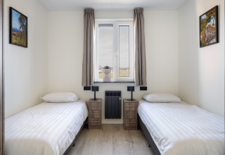 Twee eenpersoonsbedden in een lichte, eenvoudige kamer met raam, schilderijen en nachtkastjes in Zandduin Deluxe, Nederland.