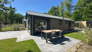 Moderne Lodge mit Esstisch und Stühlen auf einer Terrasse, umgeben von grünem Rasen und Bäumen.