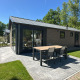 Moderne lodge med udendørs spisebord og stole på en flisebelagt terrasse, omgivet af grønne træer.