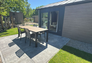 Terras bij lodge in de zon met tafel, vier stoelen op tegels, schuifdeur en verzorgd gazon.