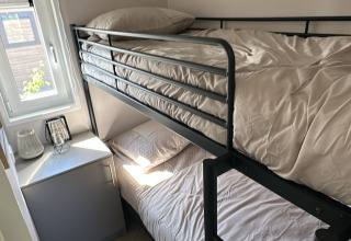 Compacte lodgekamer met metalen stapelbed, raam, klein tafeltje en minikoelkast in daglicht.