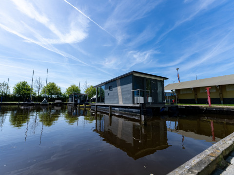 Casa flotante moderna en el agua en Marina Heeg, Países Bajos, rodeada de cielo azul y árboles verdes.