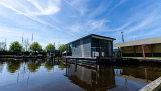Modernes Hausboot am Wasser bei Marina Heeg in den Niederlanden, mit blauem Himmel und grünen Bäumen.