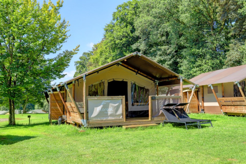 Luxury tent with wooden deck and loungers on green grass at Camping La Colline, Belgium, surrounded by trees.