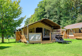 Luxury tent with wooden deck and loungers on green grass at Camping La Colline, Belgium, surrounded by trees.