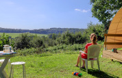 Kvinde sidder på stol foran telt, nyder udsigt over grønne marker på Camping La Colline, Belgien.