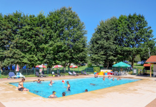 Bambini e adulti si divertono nella piscina all'aperto del Camping La Colline, circondata da ombrelloni e alberi.