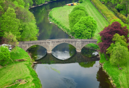 Vue aérienne d'un pont en pierre traversant une rivière, espaces verts et hébergements glamping dans un parc de vacances.