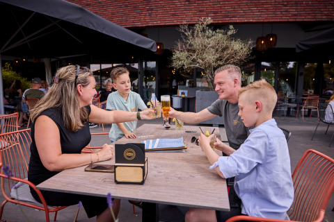 Gezin geniet van een maaltijd op het terras van vakantiepark De Leistert in Limburg, Nederland, in de zon.