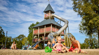 Børn leger i sandet foran en stor legeplads med rutsjebane i ferieparken De Leistert, Limburg, Holland.