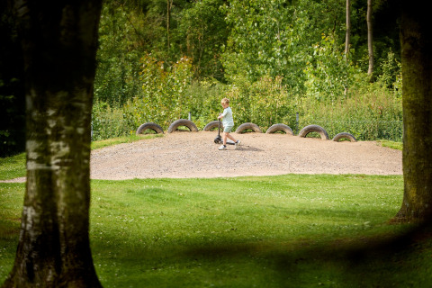 Kind fährt Roller auf einem Sandhügel mit alten Reifen im De Leistert Ferienpark, Limburg, Niederlande.