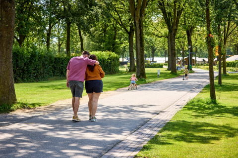 Par i De Leistert-ferieparken i Limburg, Holland, går på en allé med børn, træer og telte i baggrunden.