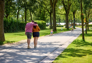 Couple walking in De Leistert holiday park, Limburg, Netherlands, with children playing and trees lining the path.