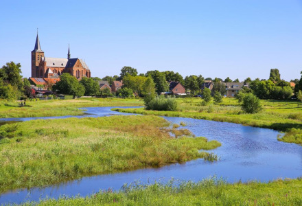 Landskab i nærheden af Roggel, Limburg, Holland, med en kirke, flod og grønne enge under blå himmel.