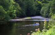 Two people kayaking on a calm river surrounded by green forest at a holiday park with glamping sites.