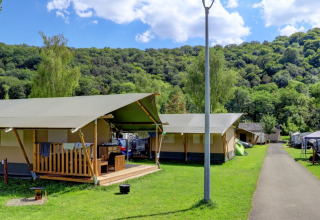 Tentes de glamping dans un parc de vacances, entourées de collines vertes, arbres et ciel bleu nuageux.