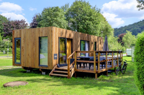 Modern wooden glamping cabin with terrace at a holiday park, surrounded by greenery and blue sky.