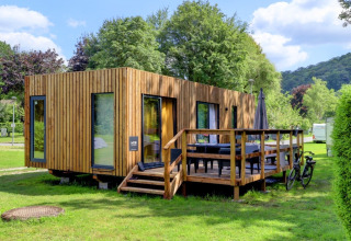 Modern wooden glamping cabin with terrace at a holiday park, surrounded by greenery and blue sky.