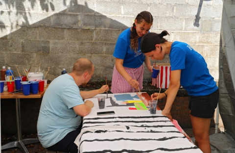 Three people painting on a large canvas outdoors at a table with art supplies at a holiday glamping park.