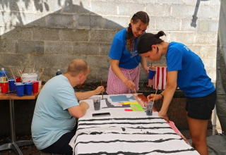 Three people painting on a large canvas outdoors at a table with art supplies at a holiday glamping park.