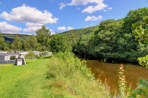 Holiday park with glamping accommodations beside a river, lush greenery, and hills under a blue sky.
