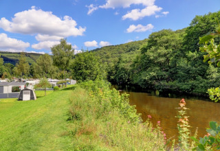Holiday park with glamping accommodations beside a river, lush greenery, and hills under a blue sky.