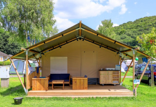 Ampia tenda glamping con veranda in legno e salottino in un villaggio vacanze immerso nel verde.