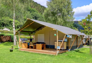 Tenda glamping in un parco vacanze con veranda in legno, circondata da alberi e colline verdi sotto il cielo blu.
