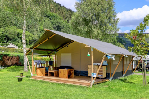 Glamping-tent op een vakantiepark met houten veranda, omgeven door bomen en groene heuvels onder blauwe lucht.