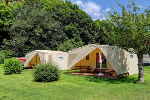 Due tende glamping di lusso con veranda in legno e tavoli da picnic in un parco vacanze verdeggiante.