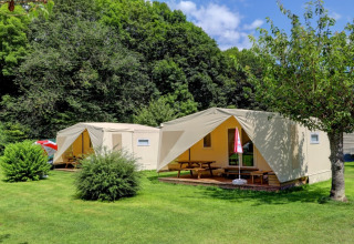 Due tende glamping di lusso con veranda in legno e tavoli da picnic in un parco vacanze verdeggiante.