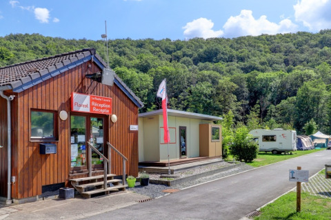 Photo of a holiday park reception, flag, caravans, and tents, nestled among green hills and trees.