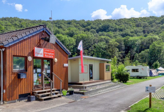 Photo of a holiday park reception, flag, caravans, and tents, nestled among green hills and trees.