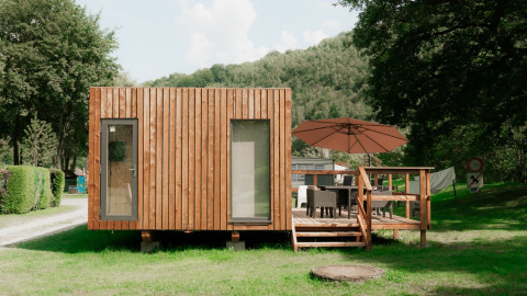 Modern wooden tiny house with a deck and umbrella, situated in a holiday park surrounded by nature.