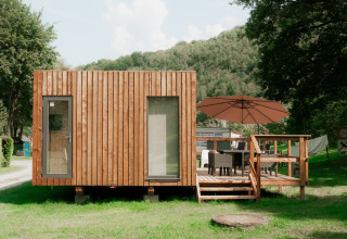 Modern wooden tiny house with a deck and umbrella, situated in a holiday park surrounded by nature.