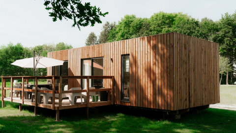 Moderne houten glampingwoning met terras, parasol en stoelen in een groene vakantiepark omgeving.
