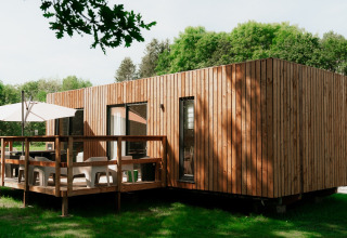 Moderna casa glamping in legno con terrazza, ombrellone e sedie, immersa in un parco vacanze verde.