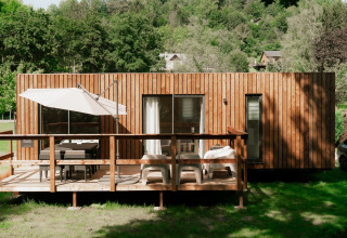 Casetta glamping in legno con terrazza, ombrellone e sedie, immersa in un parco vacanze verdeggiante.