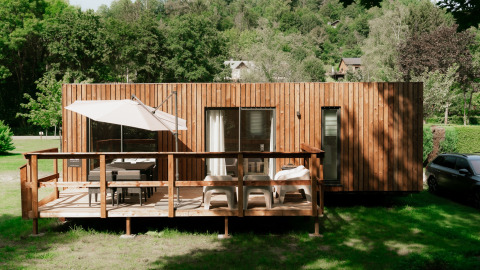 Cabaña glamping de madera con terraza, sombrilla y muebles de exterior en parque vacacional rodeado de verde.