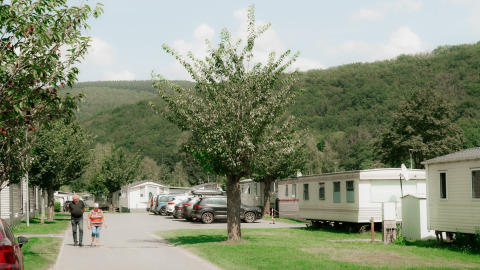 Twee personen wandelen tussen glampingwoningen in een vakantiepark met groene heuvels op de achtergrond.
