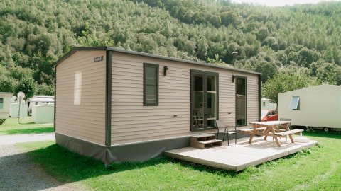Et moderne glamping-mobilhome med terrasse og bænk i en naturskøn feriepark omgivet af grønne bakker.