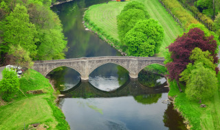 Luftaufnahme einer Steinbrücke über einen ruhigen Fluss in einem grünen Ferienpark mit Glampingzelten.