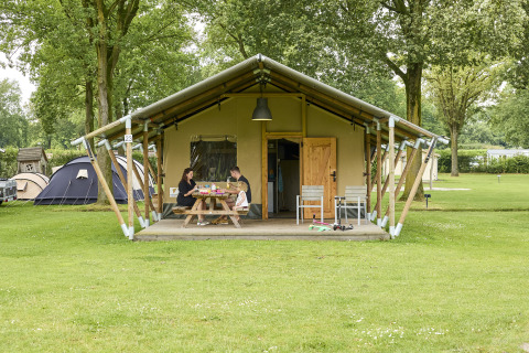 Familie frühstückt vor einem Safari-Zelt auf einem Campingplatz mit grüner Wiese und Bäumen im Hintergrund.