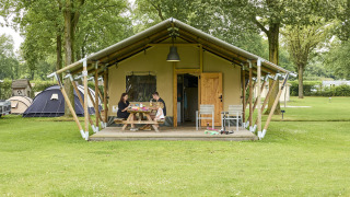 Familia desayunando en el porche de una tienda safari en un camping, rodeados de césped y otras tiendas.