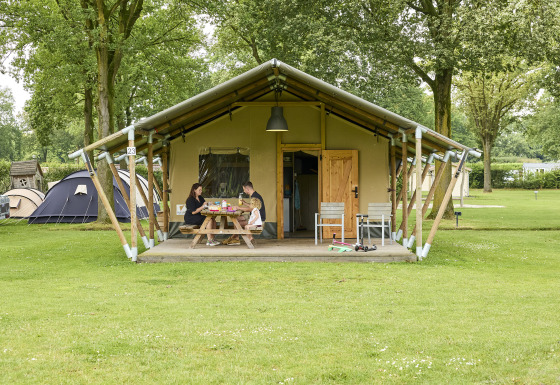 Gezin eet ontbijt voor een safaritent op een camping, omgeven door grasvelden en verschillende tenten.