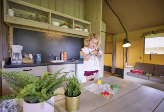 Niña usando un móvil dentro de la Safari Tent en De Leistert, Países Bajos, con cocina y juego de mesa al fondo.