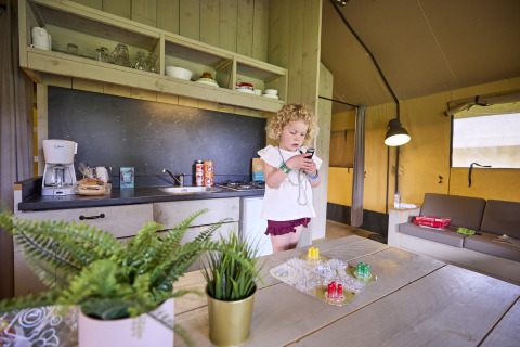 Kind speelt met smartphone in de Safari Tent bij De Leistert, Nederland, met keuken en bordspel op tafel.