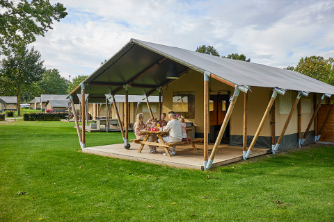 Familie hygger sig foran en Glamping Tent Comfort safari-telt på en grøn campingplads under åben himmel.